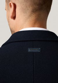 Granatowy wełniany płaszcz o fakturowanym wykończeniu, z naszywką z napisem "PROCHNIK" na tylnej części kołnierza. Prosty, dopasowany fason.