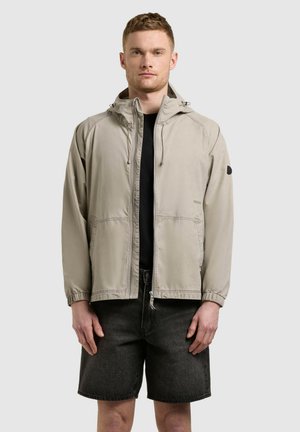 Mees, kes kannab beežikat kapuutsiga lukuga jope, musta särki ja musti teksapükse, seisab otse kaamera poole lihtsa tausta ees.