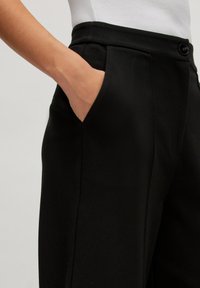 Pantalons noirs avec une taille plate, des poches latérales et une coupe ajustée. Le tissu a une texture lisse et un éclat subtil.
