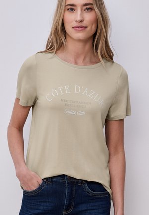 Mujer rubia con camiseta beige con el texto "Côte d'Azur Mediterranean Experience Sailing Club", combinada con vaqueros azul oscuro, con la mano en el bolsillo.
