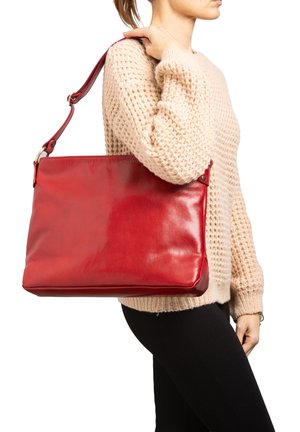 Vrouw draagt een beige gebreide trui en zwarte broek en heeft een grote rode leren schoudertas over haar rechter schouder hangen.