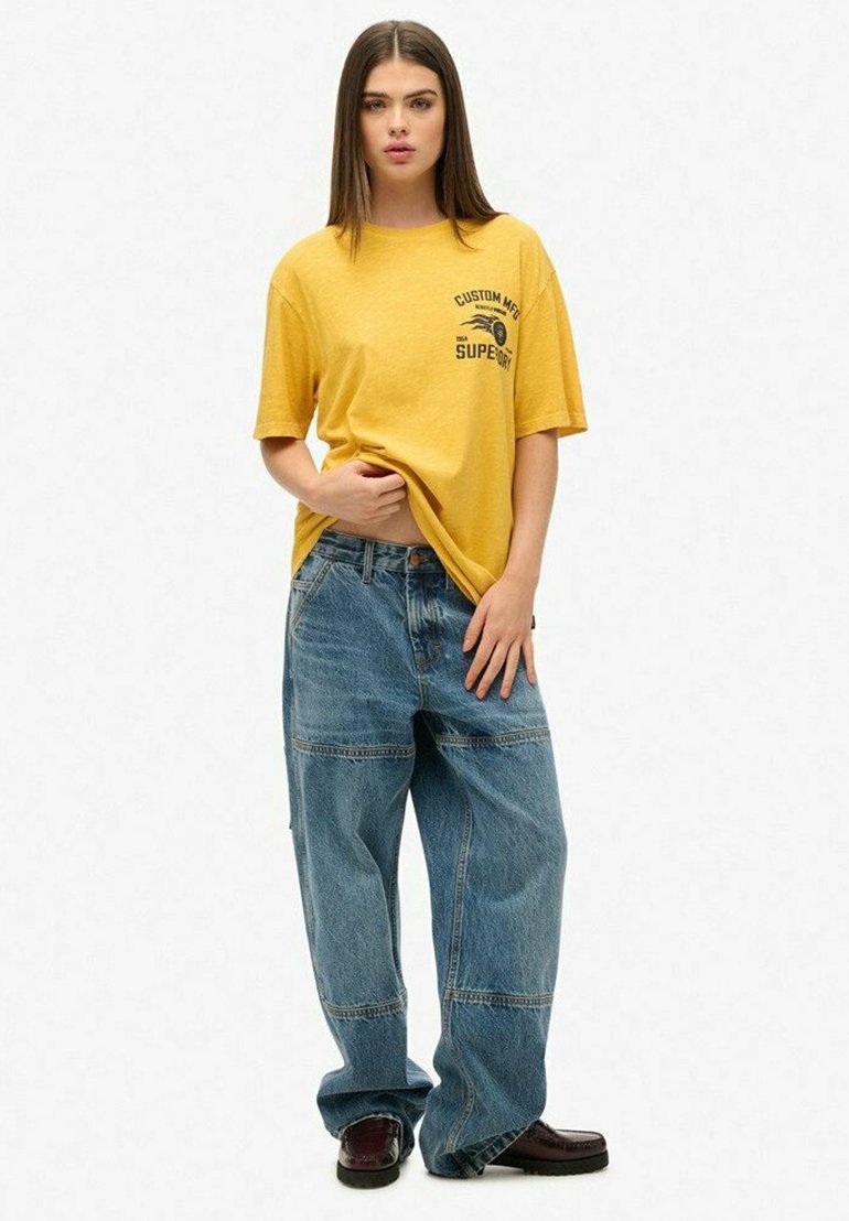 Gelbes Baumwoll-T-Shirt mit schwarzem Grafikdruck, kombiniert mit locker sitzenden blauen Jeans aus Denim mit sichtbaren Nähten und Taschen.