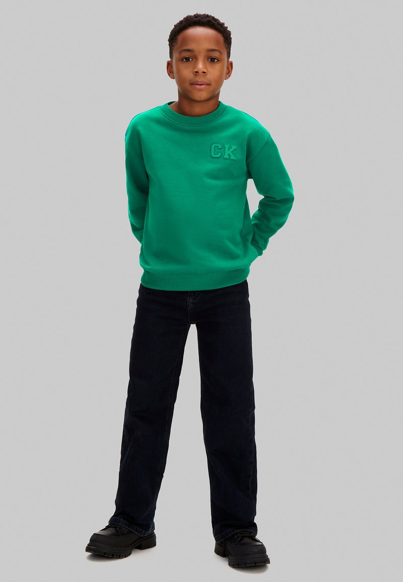 Chłopiec stojący z rękami za plecami, ubrany w zieloną bluzę z kapturem z logo "CK", czarne spodnie i czarne buty na szarym tle.