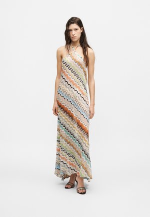 Ung kvinde iført en ærmeløs, flerfarvet maxikjole med bølgede striber og sorte åbne hæle, står mod en ensfarvet baggrund.