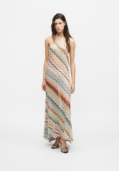 Jeune femme portant une robe maxi sans manches à rayures ondulées multicolores et des talons ouverts noirs, debout devant un fond uni.