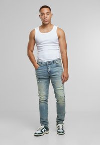 Joven con cabello corto que lleva una camiseta sin mangas blanca, jeans claros desgastados y zapatillas blancas y verdes, de pie con una mano en el bolsillo.