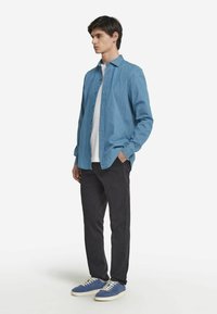 Ljusblå skjorta med knappar i en avslappnad passform, bärs över en vit t-shirt, kombinerad med svarta byxor och blå sneakers.