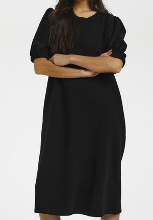 Femme aux longs cheveux foncés portant une robe noire ample longueur genoux avec des manches courtes bouffantes, bras croisés devant un fond blanc uni.
