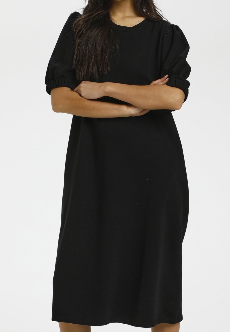 Femme aux longs cheveux foncés portant une robe noire ample longueur genoux avec des manches courtes bouffantes, bras croisés devant un fond blanc uni.