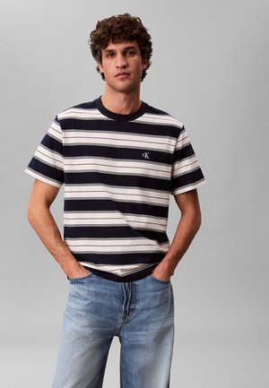 Junger Mann mit lockigem Haar, der ein schwarz-weiß gestreiftes kurzärmeliges Hemd und hellblaue Jeans trägt, Hände in den Taschen, neutraler grauer Hintergrund.