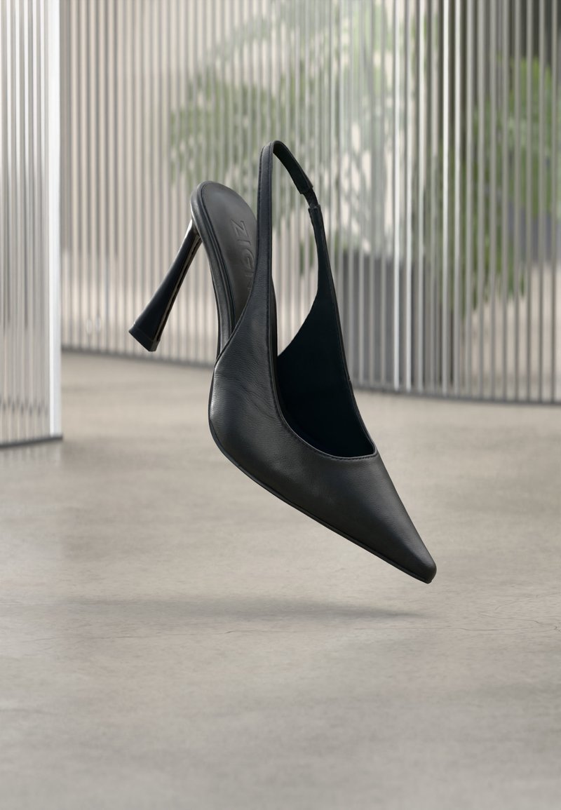 Chaussure à talon aiguille en cuir noir avec bride arrière et bout pointu, flottant au-dessus d'un sol en béton lisse et d'un panneau métallique vertical en arrière-plan.
