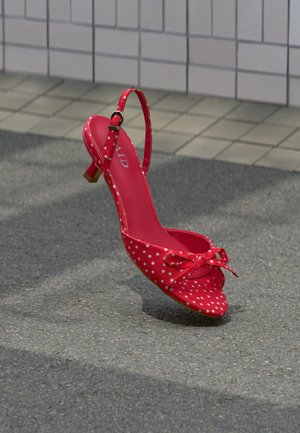 Chaussure à talon haut rouge à pois blancs avec bride à la cheville et nœud, placée verticalement sur un asphalt gris près d'un mur carrelé.