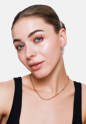 Goldene Kette, glatte Textur, schichtedesign. Modell mit zurückgebundenem Haar, blauen Augen, trägt ein schwarzes ärmelloses Top.