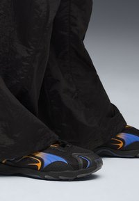 Pantalones deportivos negros con un acabado texturizado y un ajuste holgado, combinados con zapatillas negras que tienen acentos en azul y naranja y un diseño de malla.