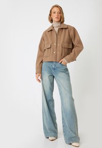 Giacca corta marrone con due tasche frontali e bottoni dorati, abbinata a jeans a gamba larga blu chiaro e un dolcevita beige.