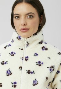 Veste en polaire avec col haut, de couleur crème, ornée de motifs floraux violets et marron, dotée d'une fermeture éclair blanche.