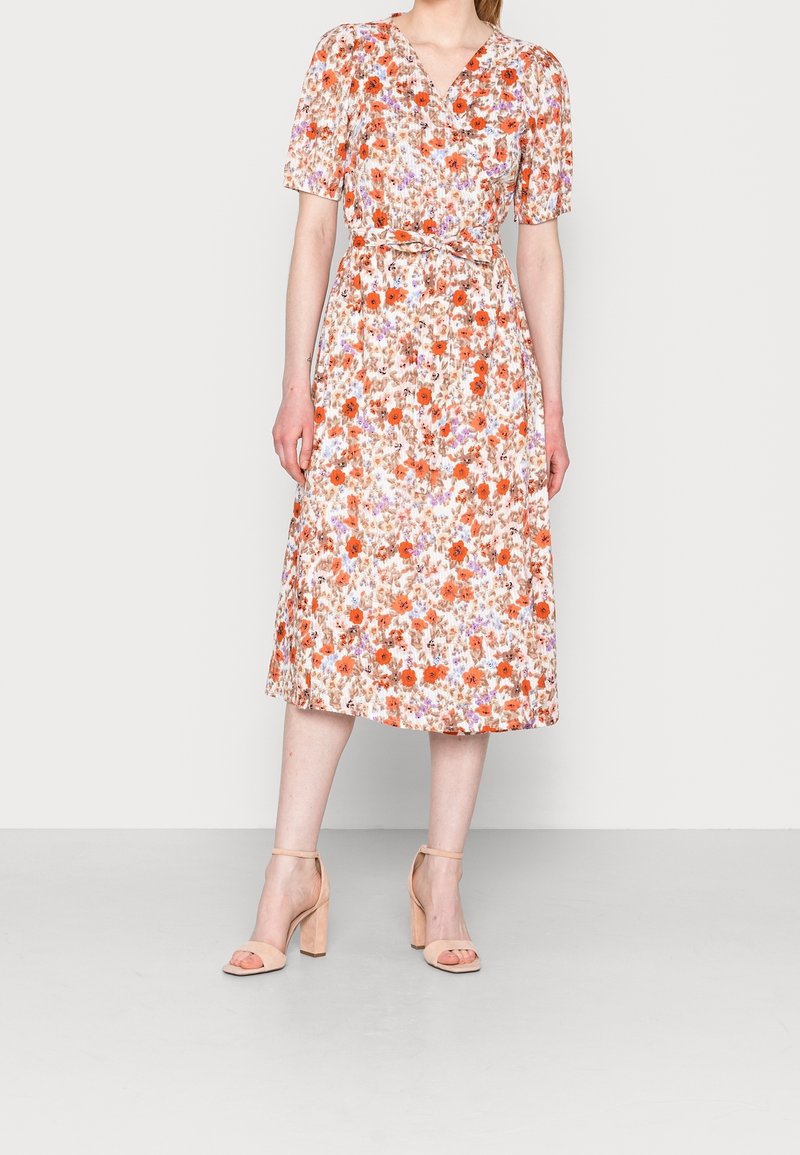 Femme portant une robe fleurie longueur genou avec des manches courtes bouffantes et des sandales à talons beiges, debout devant un fond uni.