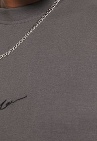 T-shirt in cotone grigio con scollatura a girocollo e tessuto testurizzato, con dettaglio ricamato in nero e una collana a catena d'argento a più strati.