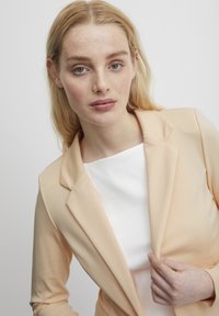 Světlý broskvový blazer s klopami, vyrobený z hladké tkaniny, spárovaný s bílým vrškem s žebrováním. Modelka drží blazer mírně otevřený.
