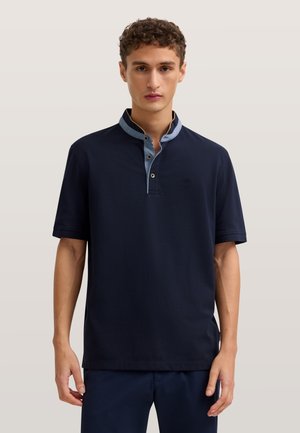 Junger Mann trägt ein dunkelblaues Hemd mit kurzen Ärmeln und einem kontrastierenden hellblauen Knopfkragen, steht vor einem einfarbigen Hintergrund.