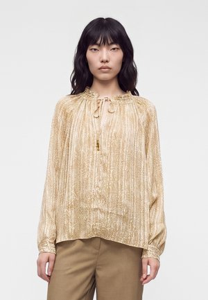 Vrouw met lang zwart haar draagt een glanzende, gestructureerde gouden blouse met een gestrikte hals en beige broek, staand tegen een effen witte achtergrond.