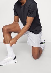 Szare sportowe szorty i czarna koszulka odprowadzająca wilgoć. Białe buty sportowe z czarnymi akcentami. Klęczy, poprawiając skarpetkę.