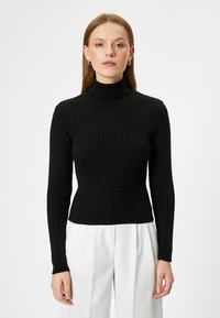 Svart ribbad polotröja med långa ärmar och en åtsittande design, tillverkad av strukturerat stickat material. Bärs med vita byxor.