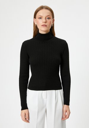 Sort ribbet turtleneck sweater med lange ærmer og en tætsiddende pasform, lavet af struktureret strikmateriale. Bæres med hvide bukser.