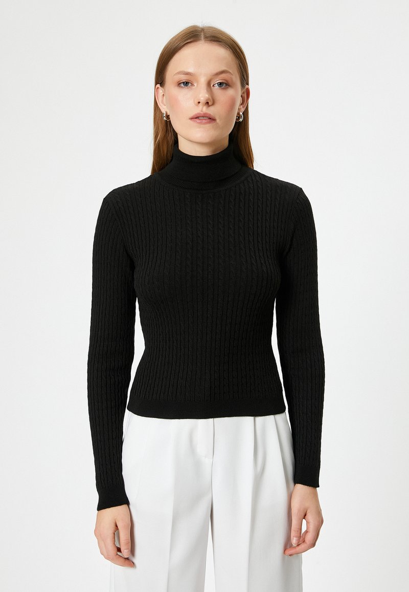 Svart ribbad polotröja med långa ärmar och en åtsittande design, tillverkad av strukturerat stickat material. Bärs med vita byxor.