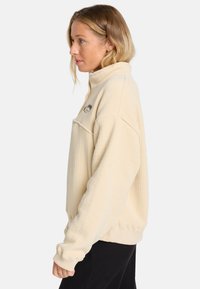 Crème ribgebreide pullover trui met een hoge kraag en een voorzak. Bevat een zwart geborduurd logo op de borst en brede mouwen.