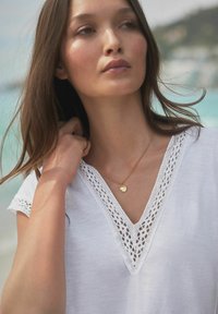 Haut blanc à manches courtes avec un col en V bordé de dentelle, associé à un délicat collier en or avec un pendentif circulaire.