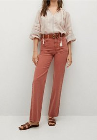 Blouse rose pâle avec broderie, taille cintrée et pampilles, assortie à un jean taille haute couleur rouille à jambes larges et des sandales marron.