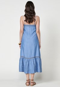 Robe midi en denim bleu clair avec bretelles spaghetti, garniture brodée, ourlet ruffle à volants et coupe décontractée. Pas de motifs ni de matériel notables.