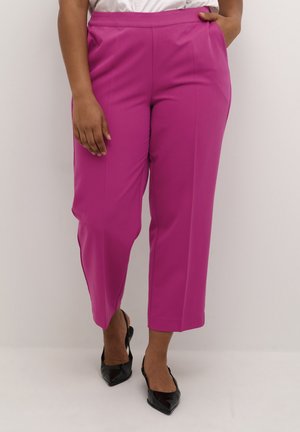 Vrouw draagt felroze korte broek en zwarte pumps met spitse neus, staand tegen een effen lichte achtergrond.