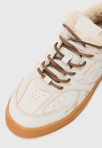 Sneaker bianca in suede con lacci marroni, dotata di un rivestimento peloso e suola di gomma beige. Accenti del logo dorato sulla linguetta e sul lato.