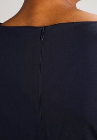 Zbliżenie na tylną część dekoltu i zamek błyskawiczny ciemnoniebieskiego ubrania na osobie o średniej brązowej skórze.