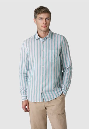 Junger Mann, der ein hellblau-weiß gestreiftes Hemd mit langen Ärmeln und beige Hosen trägt, steht vor einem schlichten grauen Hintergrund.