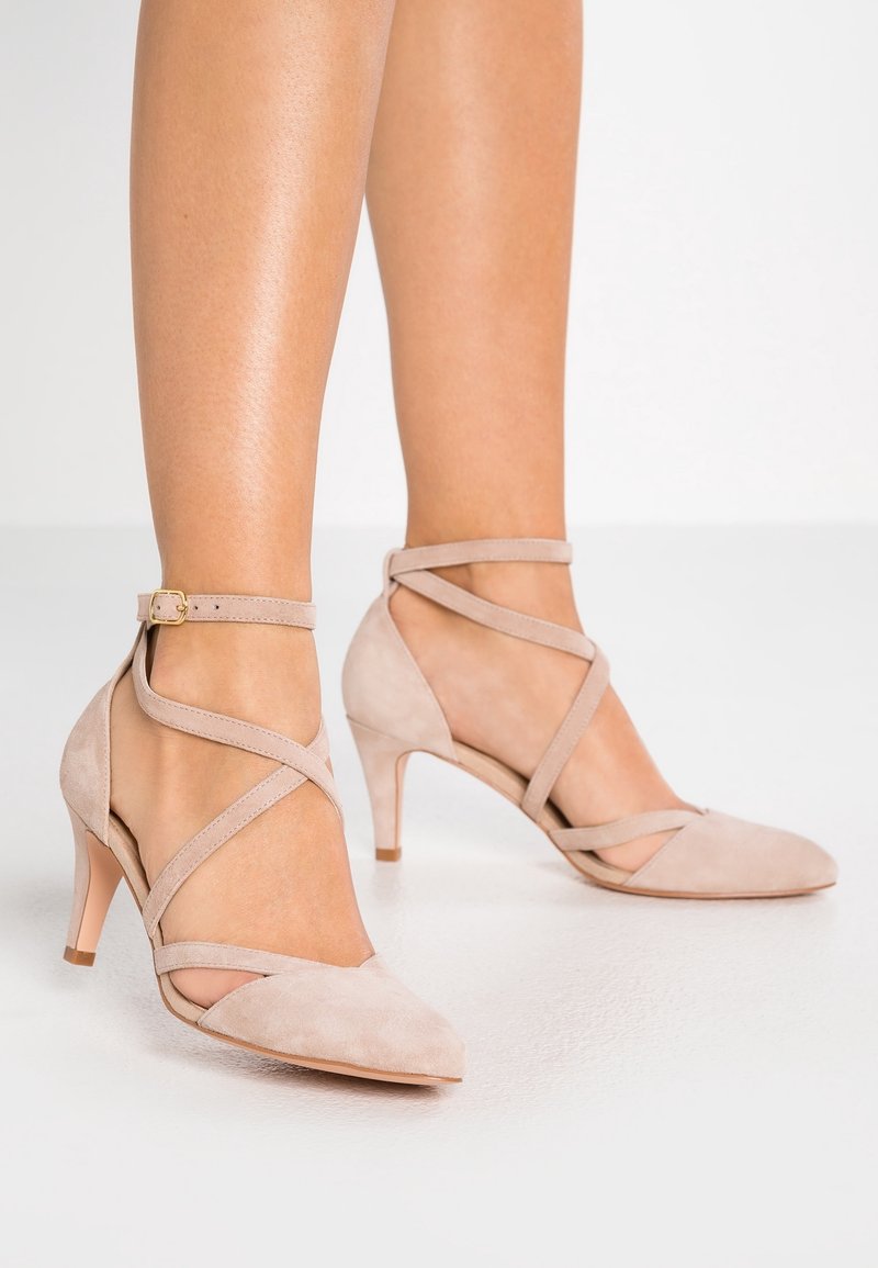 Pieds chaussés d'escarpins beige en daim à bout pointu avec des brides croisées à la cheville et une petite boucle, debout sur un sol blanc.