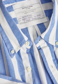 Camicia in cotone a righe blu e bianche con colletto button-down, bottoni bianchi e un'etichetta di cura che indica "DEADSTOCK" e dettagli sul recupero.