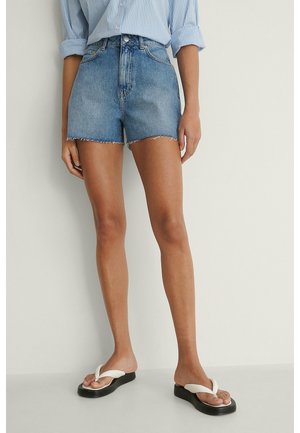 Person iført højttaljede denimshorts, en blåstribet skjorte med op-rullede ærmer og hvide platform flip-flops, stående mod en hvid væg.
