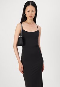 Črna oprijeta obleka z ramenskimi trakovi, gladka tekstura in subtilen lesk; model nosi majhno črno torbico s verižnim ročajem.