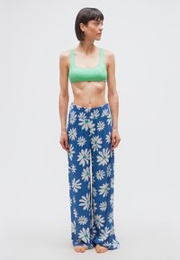 Γυναίκα που στέκεται με κοντά σκούρα μαλλιά, φοράει πράσινο crop top και μπλε παντελόνι με φαρδύ πόδι με μεγάλα λευκά σχέδια μαργαρίτας, ξυπόλυτη σε απλό φόντο.