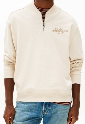 Beżowa bluza z suwakiem do ćwiartki i haftem "Tommy Hilfiger" na piersi, noszona na paskowanej koszuli i zestawiona z niebieskimi dżinsami.