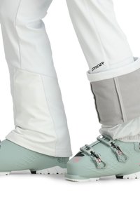 Ski-Stiefel in mintgrün mit strukturierten Seiten und weißer Sohle, ausgestattet mit metallischen Schnallen. Kombiniert mit weißen Schneehosen und grauem Fleece-Bündchen.