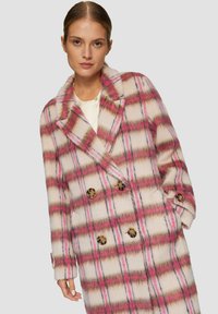 Vrouw in een dubbelzijdige crèmekleurige jas met een roze en bruine ruitpatroon en grote schildpadknopen, poseert met haar hand in haar zak.