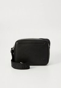 Schwarze Leder-Umhängetasche mit glatter Textur, rechteckiger Form, Reißverschluss und verstellbarem Riemen mit "Calvin Klein"-Logo.