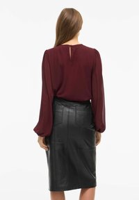 Camicetta bordeaux con maniche a sbuffo e dettaglio a chiave sulla schiena, abbinata a una gonna a tubino in pelle nera aderente con accenti delle cuciture.