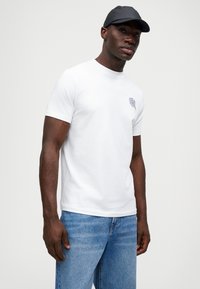 T-shirt branca de manga curta com decote redondo, apresentando uma pequena gráfica cinza no peito. Combinada com jeans de denim azul claro e um boné escuro.