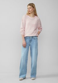 Lichtroze gebreide trui met een gestructureerd patroon, gecombineerd met lichtblauwe wijde jeans. Model staat met één hand op de heup, en toont de outfit.