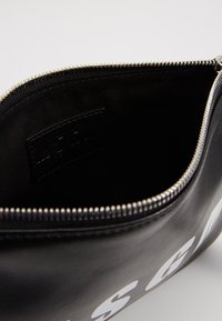 Pochette en cuir noir avec fermeture éclair, ornée de lettres blanches à l'extérieur et avec un intérieur doublé de tissu visible par la fermeture éclair ouverte.